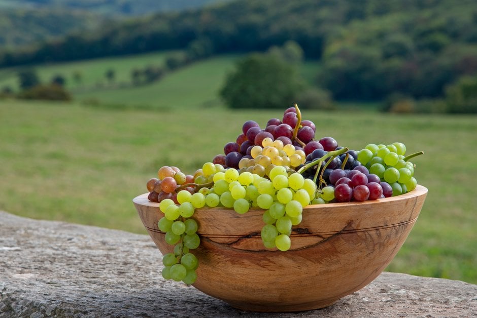 A wooden bowl of mixed dessert grapes with a countryside backdrop